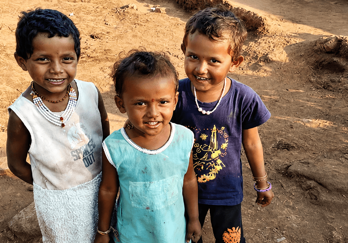 Drei Kinder stehen lächelnd auf einem staubigen Dorfplatz in Indien und blicken in die Kamera.