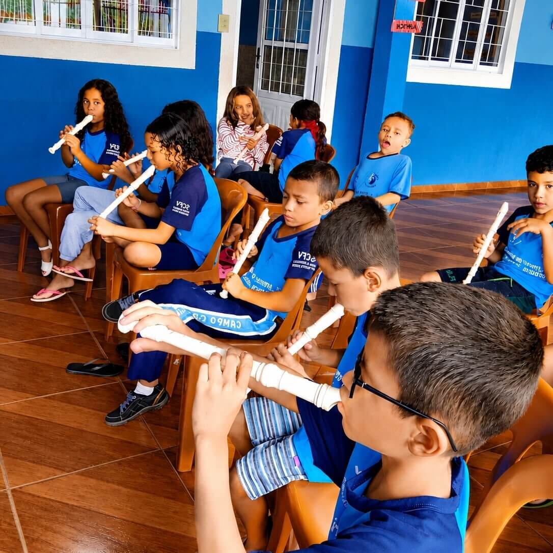 Mehrere Kinder in blauen T-Shirts sitzen auf Stühlen in einem Schulflur und spielen gemeinsam auf weißen Blockflöten. Einige üben konzentriert, während andere aufmerksam zuhören.