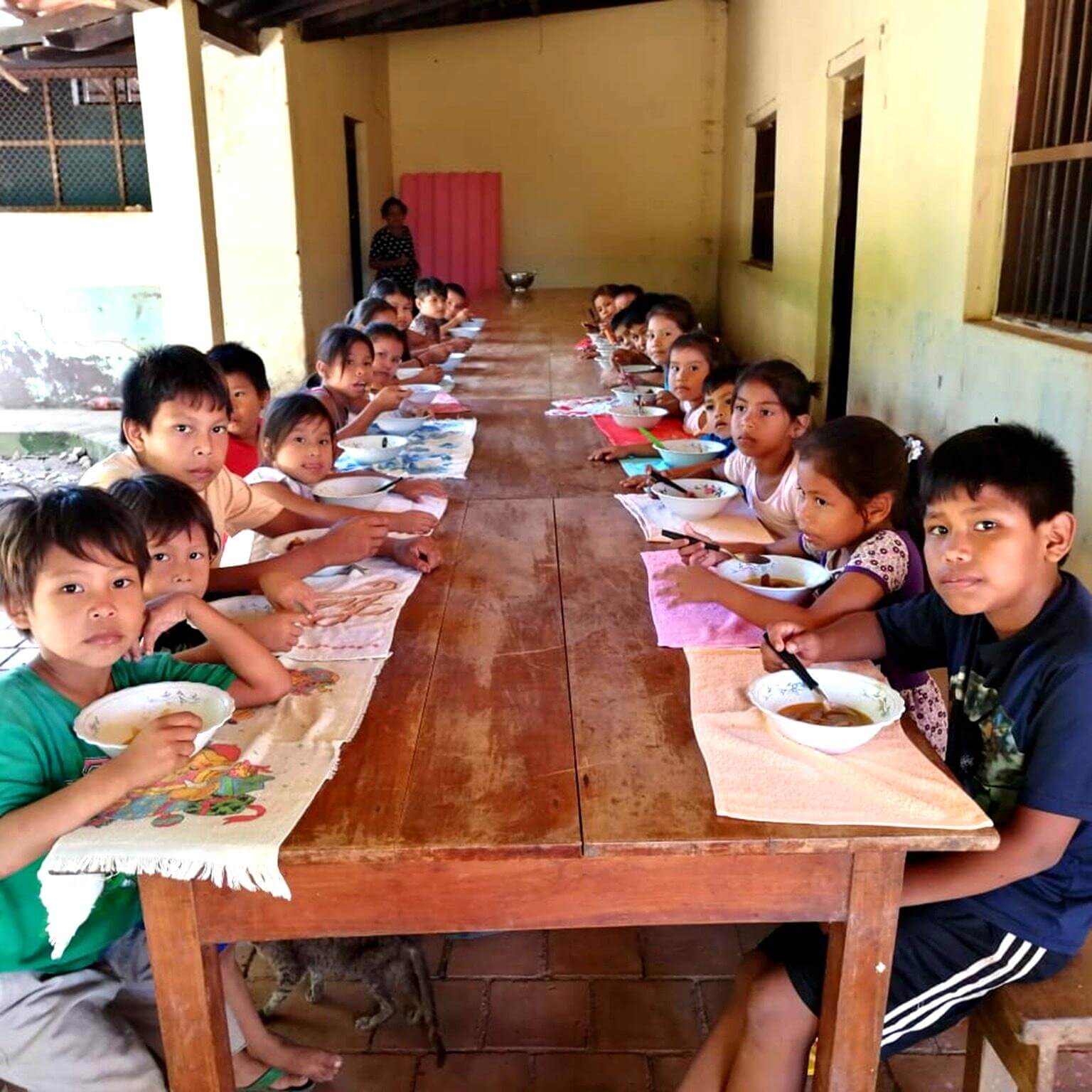 Viele Kinder sitzen an einem langen Holztisch unter einer Veranda und essen aus Schüsseln; die meisten schauen zur Kamera, während am Ende des Tisches eine erwachsene Person steht.