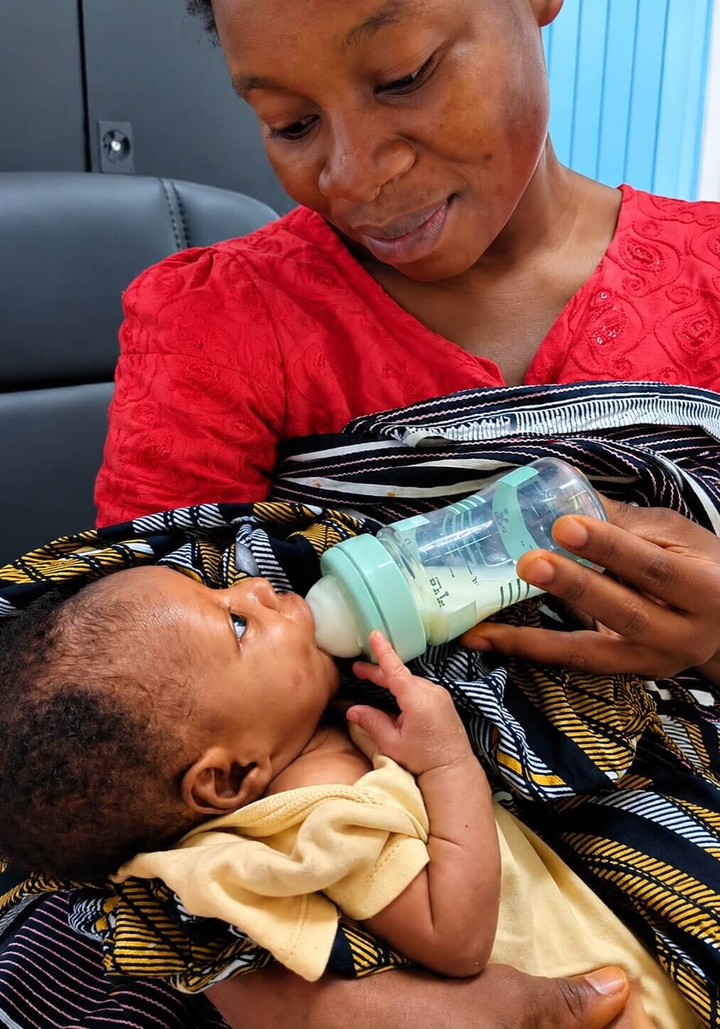 ine Mutter in einem roten Oberteil gibt ihrem Säugling liebevoll die Flasche. Das Baby liegt in ihren Armen und trinkt Milch, während die Mutter lächelnd auf ihr Kind blickt.