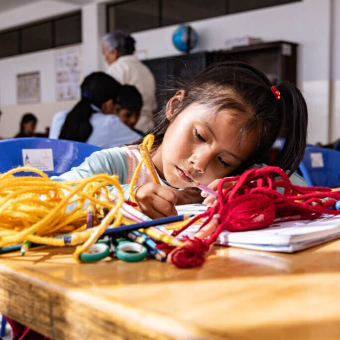 Ein Mädchen sitzt in einem Klassenzimmer an einem Tisch und malt konzentriert in ein Heft; vor ihr liegen bunte Wollfäden, Stifte und eine Schere, im Hintergrund arbeiten weitere Kinder.