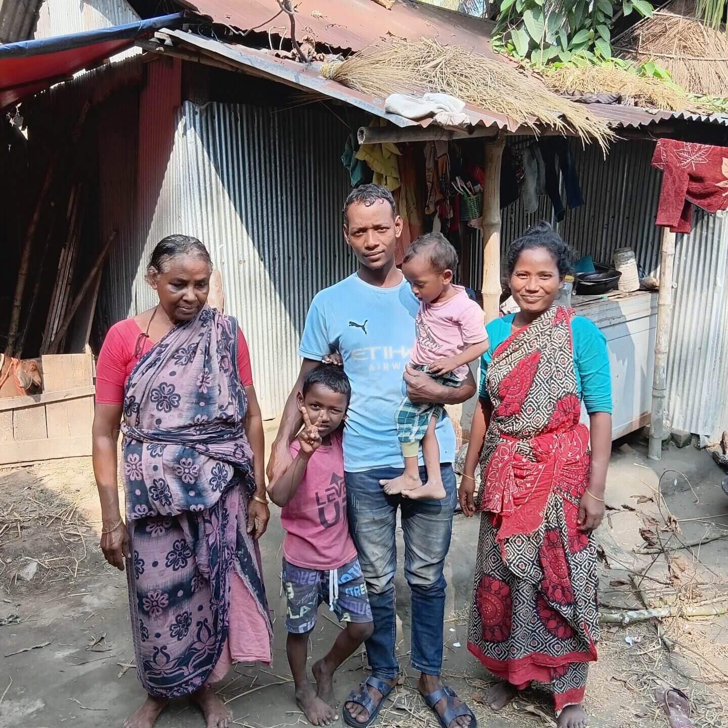 Fünf Personen – drei Erwachsene und zwei Kinder – stehen eng beieinander vor einem einfachen Haus aus Wellblech und Holz; eines der Kinder wird auf dem Arm getragen, alle blicken zur Kamera.