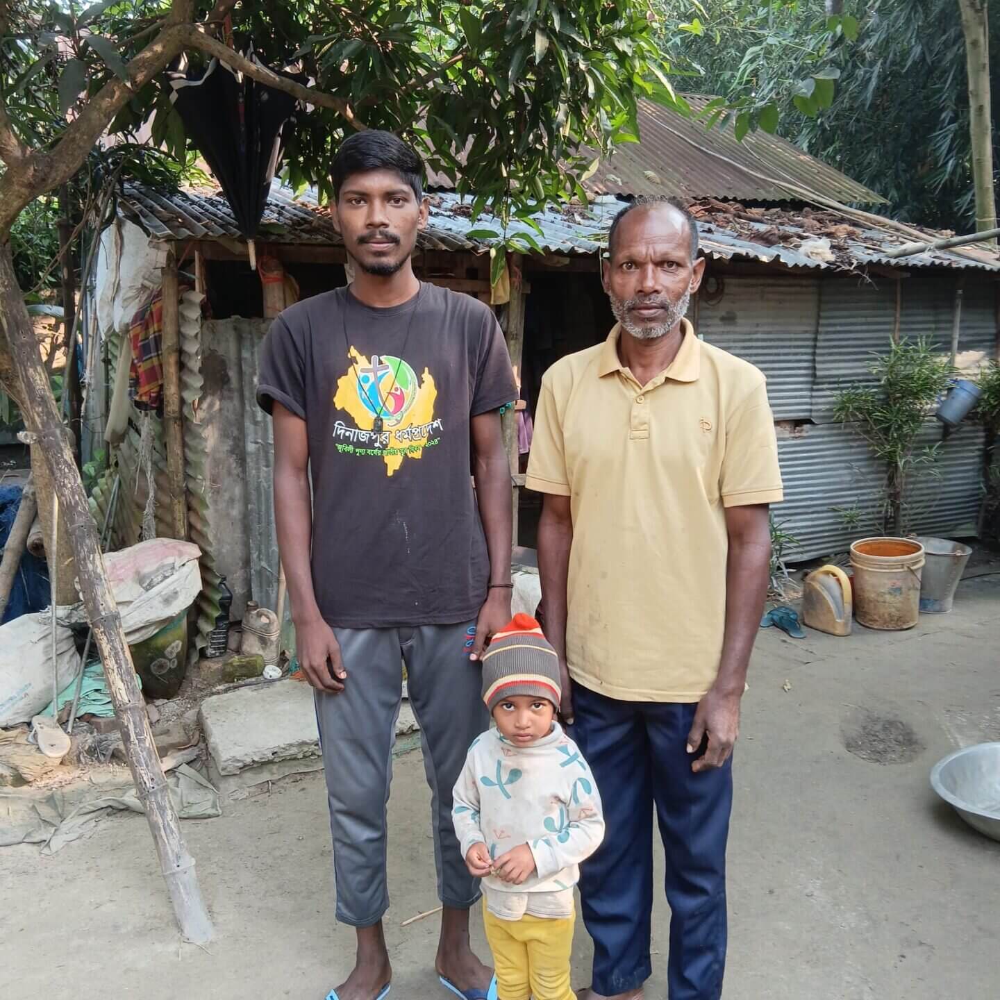 Drei Personen stehen im Freien vor einer Hütte aus Wellblech: zwei erwachsene Männer und ein kleines Kind in der Mitte; sie blicken zur Kamera, umgeben von Bäumen und einfachen Alltagsgegenständen.
