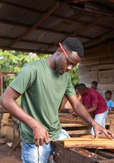 Ein junger Mann arbeitet konzentriert an einem Holzbrett in einer einfachen Werkstatt; im Hintergrund sind weitere Personen bei handwerklicher Arbeit mit Holz zu sehen.
